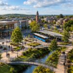Modern town centre with landscaped waterways, pedestrian bridges, green spaces, public transport, and contemporary buildings under a bright blue sky.