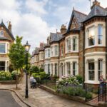 Row of Victorian houses on a quiet residential street with ornate brick facades, bay windows, flower-filled gardens, pedestrians walking and chatting, and a clear sky above.