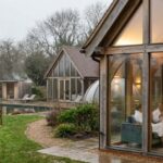 Glass house retreat in a countryside setting with modern cabins, people relaxing indoors, and a peaceful natural landscape with a pond and greenery.