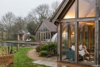 Glass house retreat in a countryside setting with modern cabins, people relaxing indoors, and a peaceful natural landscape with a pond and greenery.