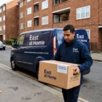 Delivery driver carrying an East at Home meal box beside a branded delivery van parked outside modern brick apartment buildings in a residential neighborhood.