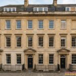 Elegant Georgian townhouses with symmetrical stone façades, tall sash windows, black front doors, and iron railings lining a quiet historic street under a clear sky.