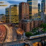 Modern built environment city skyline at dusk featuring high-rise buildings, a busy highway, railway tracks, and urban infrastructure with illuminated skyscrapers and heavy traffic.
