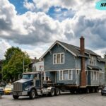 A large house being transported on a truck through a neighborhood, illustrating Sky Moving Home, home relocation, house moving, and professional moving services under a dramatic cloudy sky.