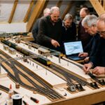 Model railway enthusiasts working on a detailed train layout in a workshop with tracks, miniature stations, tools, and a laptop displaying a railway control diagram.