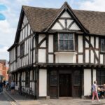 Historic The Old Crown Public House, a timber-framed Tudor-style building on a lively street with pedestrians, outdoor seating, and colorful murals in the background.