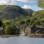 "Peaceful lakeside village with stone houses, boats on the water, and green hills rising in the background under a partly cloudy sky."
