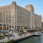 New Mart Building along the Chicago River with boats, a riverside walkway, and the city skyline.