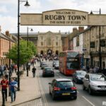 Busy street in Rugby Town featuring the iconic welcome sign, historic buildings, pedestrians, cars, and a red double-decker bus in the lively town centre.