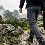 Two hikers wearing nylon pants walk along a rocky mountain trail surrounded by misty peaks and alpine vegetation.
