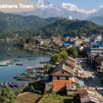 Aerial view of Pokhara Town beside a tranquil lake, colorful boats, hillside buildings, lush greenery, and snow-capped Himalayan mountains.