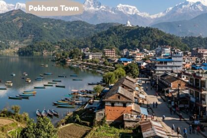 Aerial view of Pokhara Town beside a tranquil lake, colorful boats, hillside buildings, lush greenery, and snow-capped Himalayan mountains.