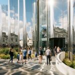 Modern mirrored artwork building with reflective panels, landscaped walkways, and pedestrians in a busy urban plaza.