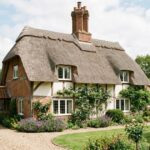 Traditional exterior view of Caston House featuring a thatched roof, brick and timber framing, a brick chimney, and a lush front garden with a gravel driveway.