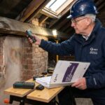 RICS certified surveyor inspecting an old property loft for a Level 3 building survey report, holding testing equipment near exposed brick walls and wooden beams.