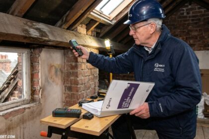 RICS certified surveyor inspecting an old property loft for a Level 3 building survey report, holding testing equipment near exposed brick walls and wooden beams.