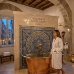 Luxury bathroom in vacation apartments Rome featuring a copper soaking tub, intricate mosaic tile wall, stone arches, and open windows overlooking a Roman street.